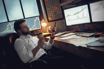 Young male trader at office work concept sitting drinking coffee browsing smartphone