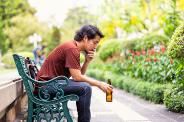 Sad single asian man drinking beer and sitting on the floor at public park.