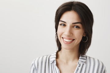 Obraz premium Attractive european shop assistant in stylish earrings and blouse smiling broadly and politely while gazing at camera, wanting to help customer to pick gift for girlfriend, standing over gray wall