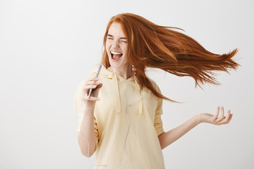 Music boosts confidence and happiness. Portrait of emotive charming woman with red hair dancing and singing song, holding smartphone like microphone, listening songs in earphones over gray background