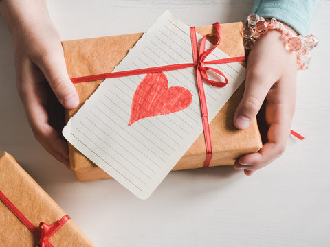 Little daughter holding a gift in a beautiful box with a red ribbon and a card with a painted heart for her beloved parents. The concept of a happy family