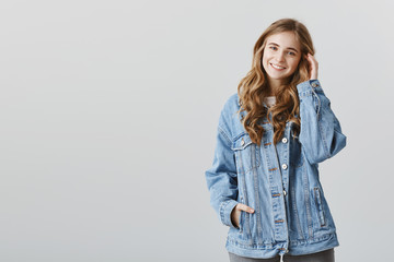 Attractive girl talking with stranger in cafe. Portrait of pleased stylish young woman in denim jacket, touching hair and smiling broadly, flirting while hanging around with hot guy over gray wall