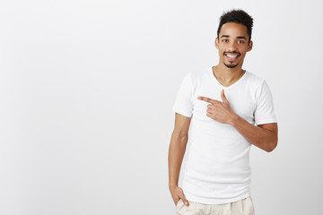 Want to show you nice place to eat. Portrait of cheerful attractive young male with afro hairstyle, smiling with approval while pointing left with index finger, suggesting great copy space