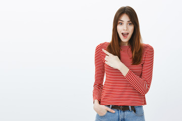 Studio shot of amazed attractive caucasian female freelancer in striped clothes, pointing at upper left corner, smiling and dropping jaw from satisfying surprising news over gray background