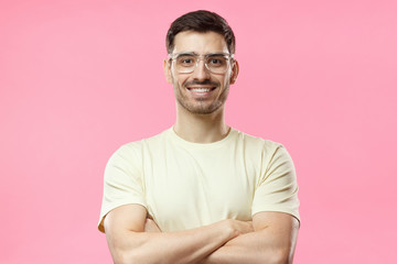 Portrait of smiling handsome man in beige tshirt and trendy transparent eyeglasses standing with crossed arms isolated on pink background