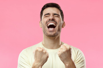 Closeup of emotional man isolated on pink background showing white teeth while screaming with closed eyes, holding hands in gesture of winner, looking extremely happy