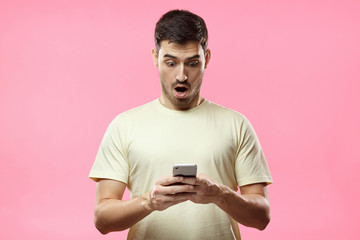 Close up portrait of handsome young man in beige t-shirt, holding smartphone, looking scared and...
