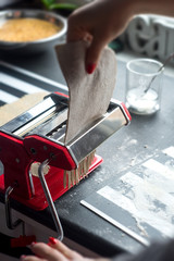 Woman rolling dough for pasta at the kitchen wooden table using red pasta grinder.