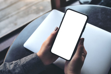 Mockup image of a woman's hand holding black mobile phone with blank white desktop screen with laptop on table in cafe
