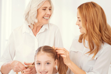 Fun and joyful moment. Selective focus on an excited beautiful elderly lady grinning broadly while talking to her mature daughter and making ponytails for her cheerful granddaughter.