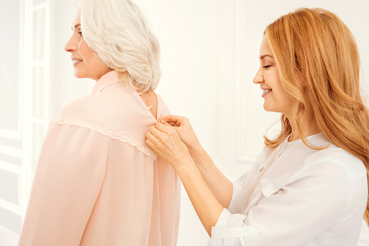Mother and daughter moment. Side view on a thoughtful adult lady standing behind her beautiful mother and helping her with buttoning a blouse while spending family time together at home.