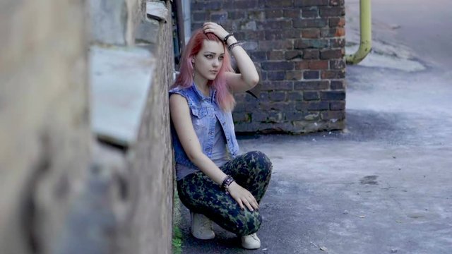Teenage girl with nose and lips piercing and pink hair sit leaning against brick wall. Slow motion. HD