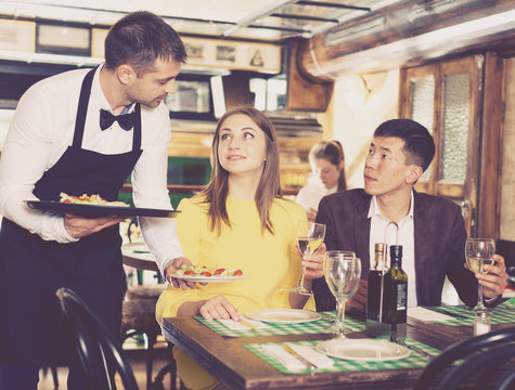 Male waiter is serves an order for smiling couple who is drinking wine and eating