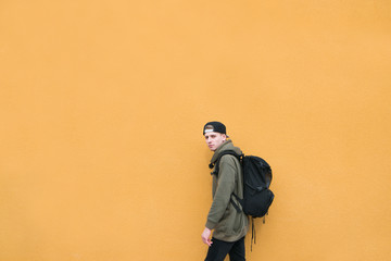 Stylish street young man with a backpack stands on the background of a large orange wall. Copyspace.