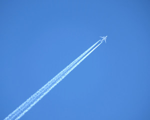 Large passenger supersonic plane flying from left to right high in blue cloudless sky and leaving long white track