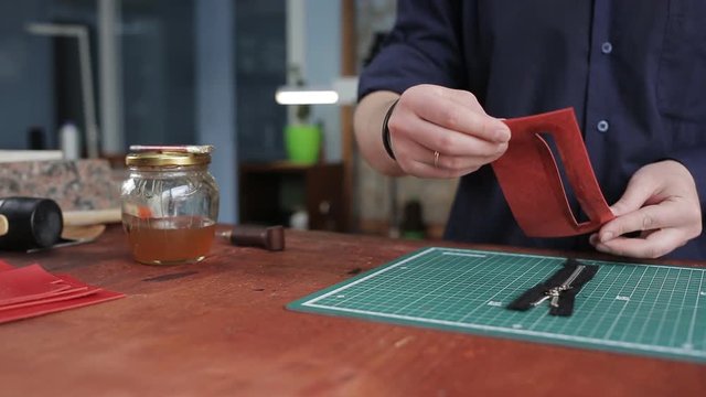 Leathersmith is applying glue on a leather parts of the zipper with a special brush. He works at the table in the workshop.