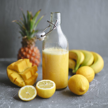 Refreshing yellow smoothie with lemons, banana, mango and pineapple on gray background