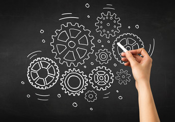 Female hand holding white chalk in front of a blackboard with gears drawn on it