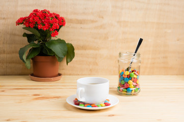 Coffee mug, red flowers, colored candy in a jar and a yellow diary on a wooden background. breakfast or lunch