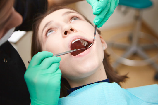 Cropped shot of dentist or hygienist in gloves checking oral cavity of young woman patient for cavities and plaque or tartar on her teeth during routing checkup, using metal probe and mirror
