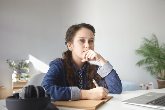 Thoughtful 12 Year Old Teenage Girl In Casual Shirt Sitting At Desk At Home With Laptop And Headphones, Doing Homework, Writing Essay In Copybook. Pensive Female Teenager Making Notes In Diary