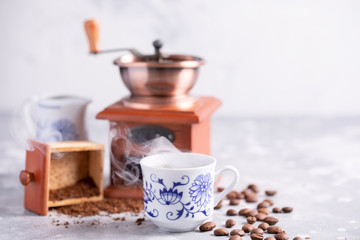 A beautiful coffee composition with a place under your text. Grains of coffee fall out of a vintage coffee grinder. Hot black coffee in a beautiful porcelain cup on the table.