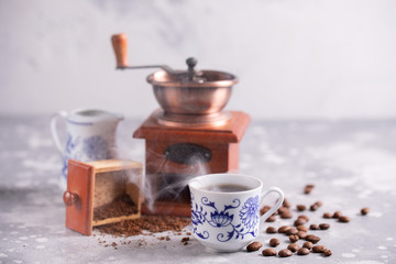 Grains of coffee fall out of a vintage coffee grinder. Hot black coffee in a beautiful porcelain cup on the table. A beautiful composition with a place under your text.