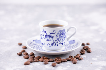 A beautiful porcelain coffee cup with hot black coffee. Coffee beans scattered around the background. Vintage china. A white-blue cup on a gray background.