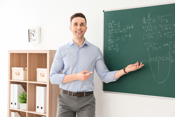 Young male teacher explaining math formulas written on blackboard in classroom