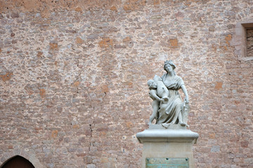 Statue on the street in front of a stone wall. A woman with a baby holding a jug.