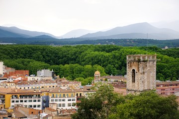 Fototapeta na wymiar View of the city of Girona from the medieval pedestrian border wall. Roofs of houses, trees, mountains in a haze in the background. Cloudy sky. Girona, Spain