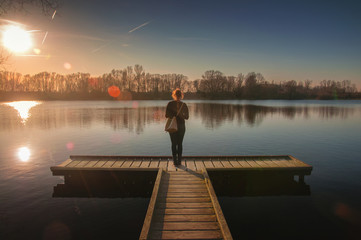 Fototapeta na wymiar Blonde girl on a wooden landing stage overlooking a lake at sunset in Braunschweig, Germany