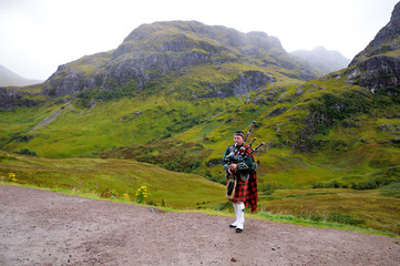 Piper in traditional Scottish outfit plays on bagpipes in Scottish Highlands. In the background of the mountain. Cloudy autumn day.