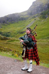 Piper in traditional Scottish outfit plays on bagpipes in Scottish Highlands. In the background of the mountain. Cloudy autumn day. - obrazy, fototapety, plakaty