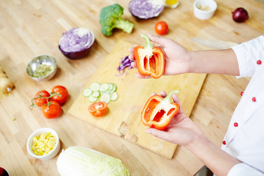 Halves of ripe red pepper on hands of chef over table with assortment of fresh vegetables for cooking