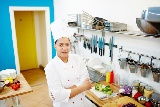 Young professional cross-armed chef looking at camera by her workplace while cooking something for clients