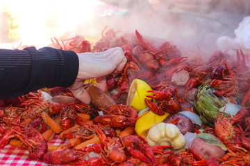 A hand reaching into a pile of steaming food piled on a table at a crawfish boil and grabbing a...
