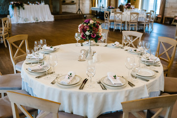 Festive table decorated with composition of flowers and greenery in the wedding banquet hall. Table covered with a tablecloth and served with dishes and cutlery. Wooden chairs. Close up.