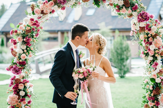 The bride and groom kissing. Newlyweds with a wedding bouquet, holding glasses of champagne standing on wedding ceremony under the arch decorated with flowers and greenery of the outdoor. - Powered by Adobe