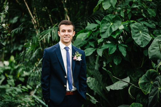 Happy handsome groom in a suit and tie is standing on the background of greenery, on the jacket is a boutonniere of flowers. Man stands in the Botanical Garden full of greenery.
