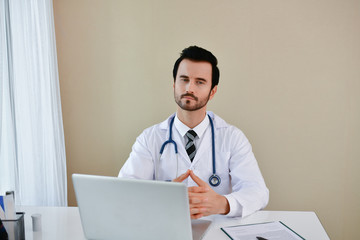 Smiling doctor posing in the office, he is wearing a stethoscope, medical staff on the hospital background