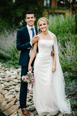 Romantic couple newlyweds, bride and groom holding bouquet of pink and purple flowers and greens, greenery with ribbon in the garden on wedding ceremony in the backyard banquet area.
