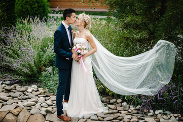 Portrait of an attractive bride who embraces and kissing the groom standing and holding bouquet of pink and purple flowers and greens with ribbon. Wedding ceremony on nature.