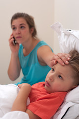 Worried young woman in a blue dress is calling to the doctor on phone on the background of sick little boy lying in bed.