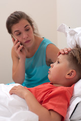 Worried young woman in a blue dress is calling to the doctor on phone on the background of sick little boy lying in bed.