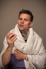 Portrait of a sick sad man with a thermometer in his mouth wrapped in a blanket on a gray background