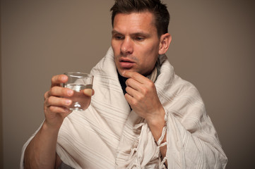 Cold and fever. Young sad sick man drinking medicine from a glass beaker during illness wrapped in a blanket on a gray background