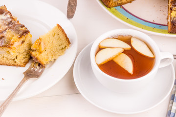 A piece of French apple cake on a plate and a cup of tea with apples on a light wooden background