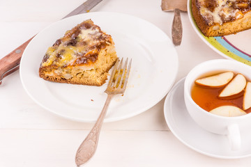 A piece of French apple cake on a plate and a cup of tea with apples on a light wooden background