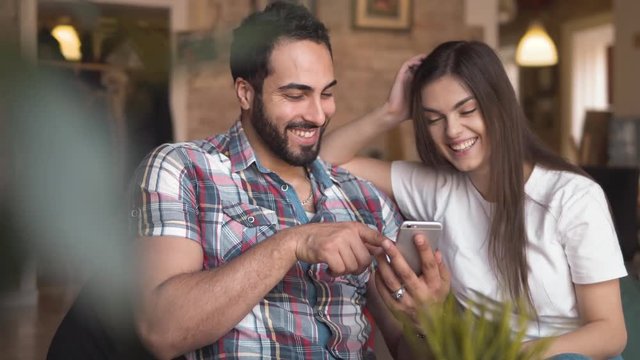 Happy young couple looking at photos on smartphone, good-looking bearded man and pretty long-haired woman enjoying sweet memories with newest technology, indoor shot in european cafe during lunchbreak